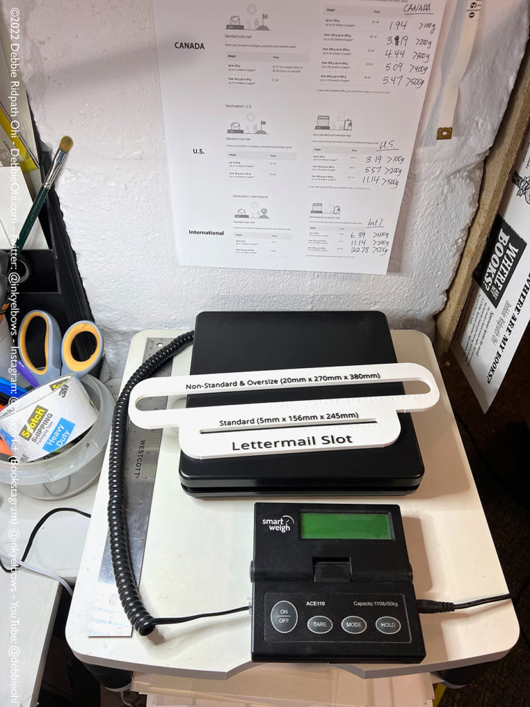 Debbie's home office package mailing set-up. Photo shows Smart Weigh scale, Canada Post slot measurement tool, packing tape, scissors. Taped up on wall: post office rates to various locations.