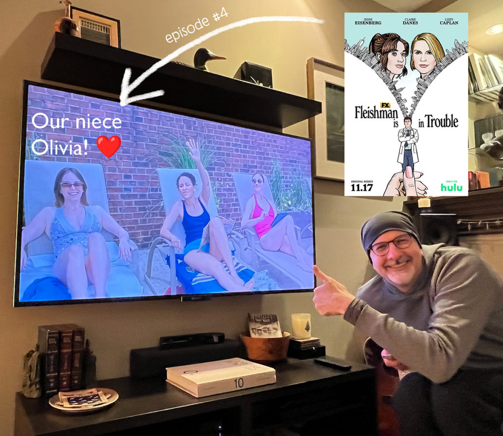 Photo of Jeff giving a thumbs up in front of our tv screen, which features a scene from Fleischman Is In Trouble with our niece as an extra. She and two others are in lounge chairs.
