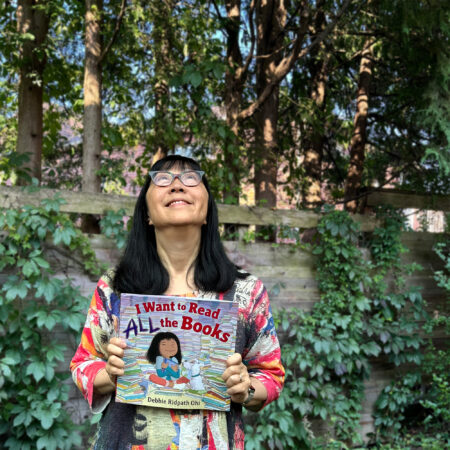 Woman holding a book called "I Want To Read ALL THe Books" looking up with a smile. In the background, is a vine-covered fence, with tall trees behind the fence.