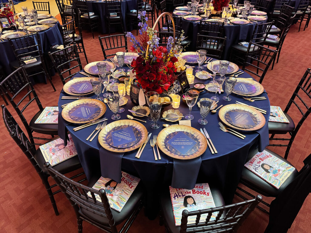 Formal round dining table covered in a dark blue table cloth and formal place settings. Each place setting has a round card with text on it. Each chair has a copy of a book called I Want To Read ALL The Books.
