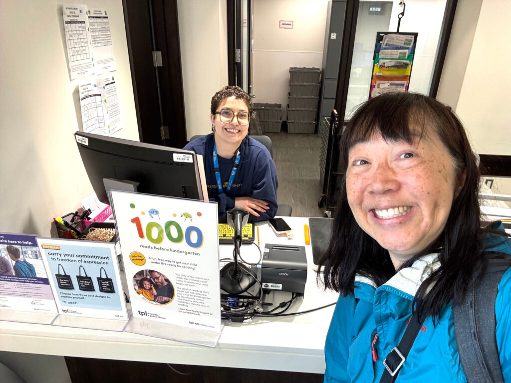 Smiling woman in front of a public library check-in desk, where another woman is seated and smiling.