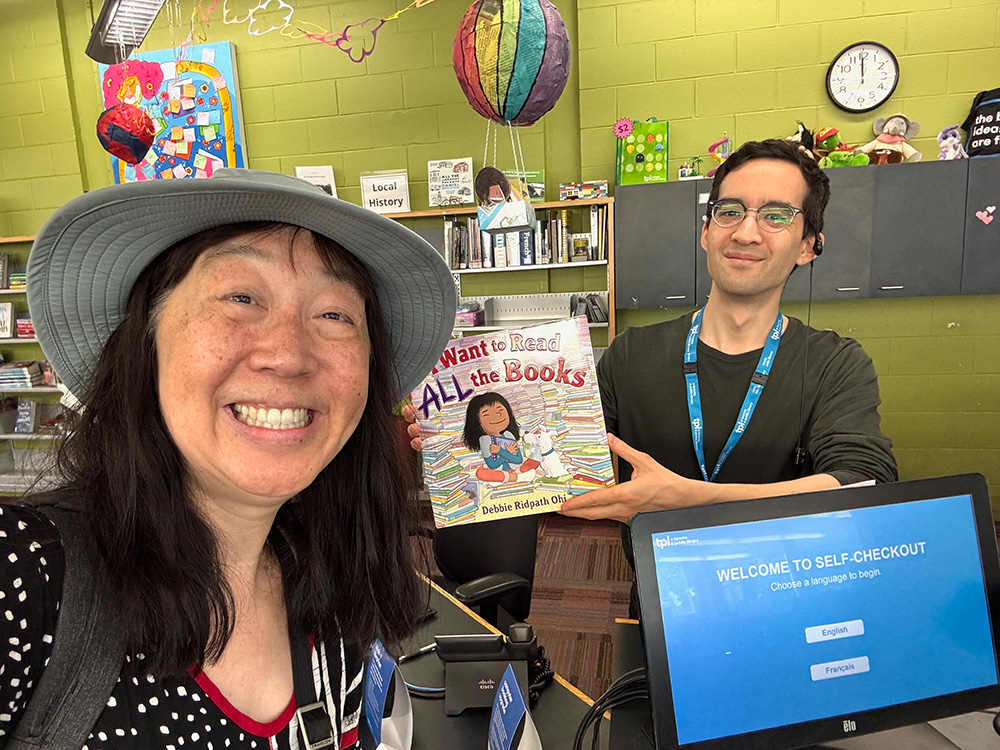 Debbie with Alex at the front desk of the Palmerston library check-out desk. Alex is holding up a copy of I Want To Read ALL The Books.