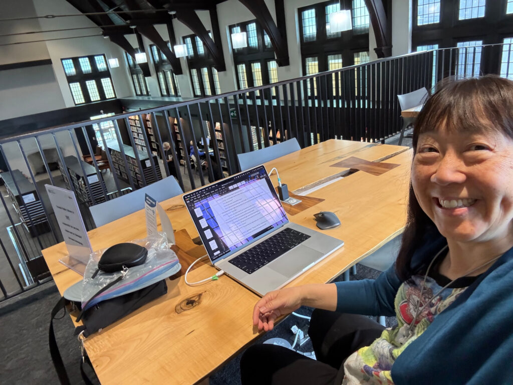 Selfie of woman at a desk with a laptop in the top floor of a library. Beyond the railing are more shelves with books.