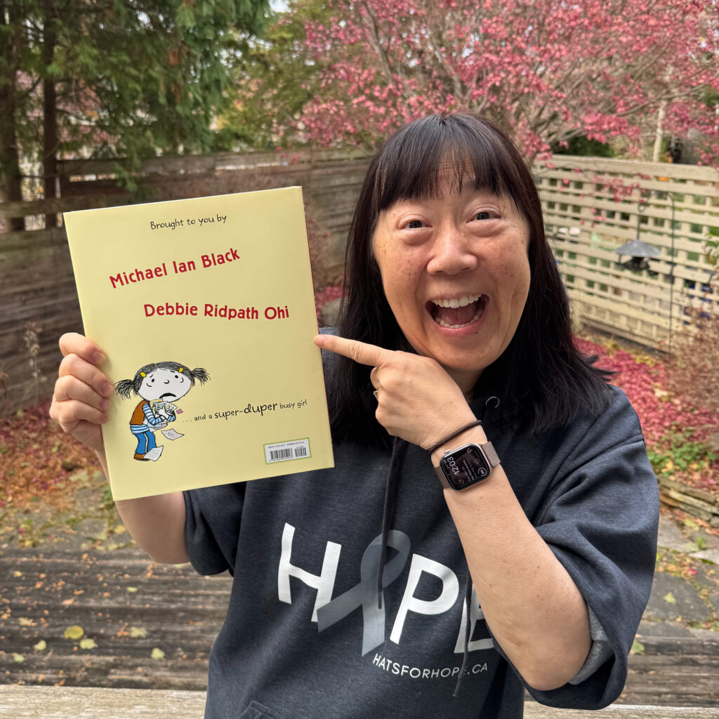Debbie showing the back cover of I'm Busy, her new picture book with Michael Ian Black, pointing to her name in the credits. Background is a backyard with fallen autumn leaves, fences, and trees.