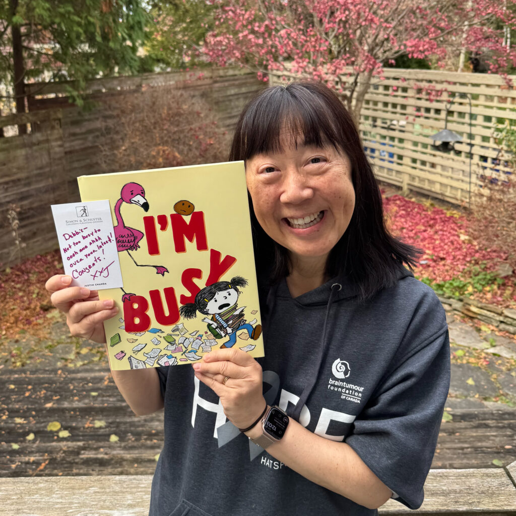 Debbie holding up I'm Busy, her new picture book with Michael Ian Black, along with a sweet note of congratulations from her editor Justin Chanda at Simon & Schuster Children's. Background is a backyard with fallen autumn leaves, fences, and trees.