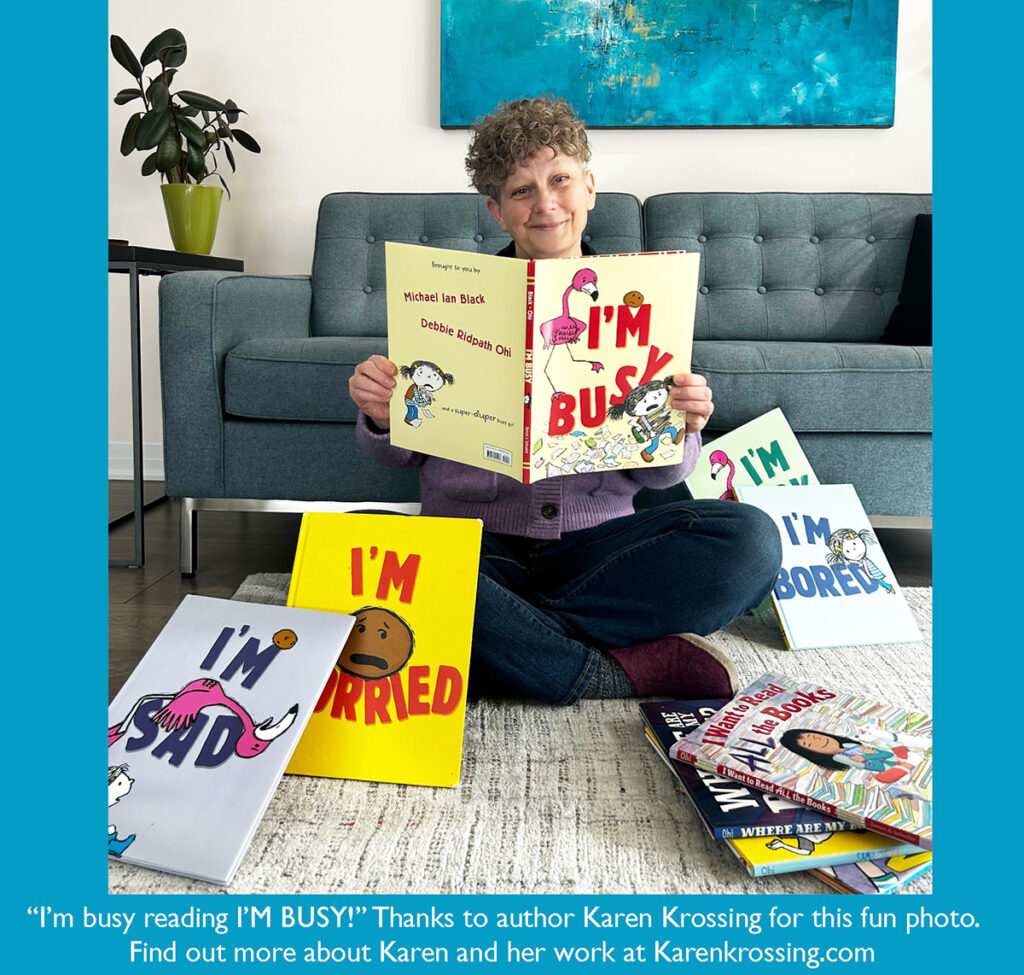 Woman sitting cross-legged on a rug in front of a gray couch, holding open the picture book I’M BUSY! by Michael Ian Black and Debbie Ridpath Ohi. Around her on the floor are other books in the series, including I’M SAD, I’M WORRIED, and I’M BORED, plus a stack of additional children’s books. Text at the bottom reads: “I’m busy reading I’M BUSY!’ Thanks to author Karen Krossing for this fun photo. Find out more about Karen and her work at Karenkrossing.com.”