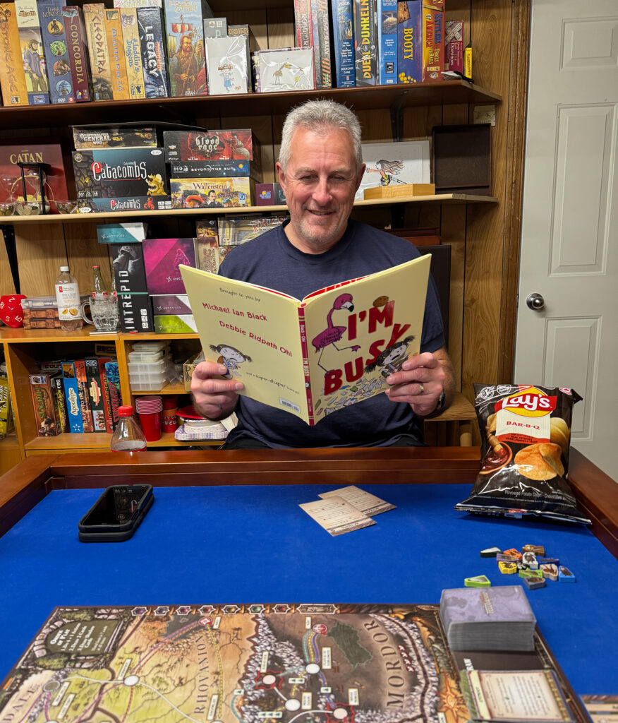Man sitting at a game table reading the picture book "I'M BUSY" by Michael Ian Black and Debbie Ridpath Ohi, with a Lord of the Rings board game set up in front of him and shelves of board games behind him.