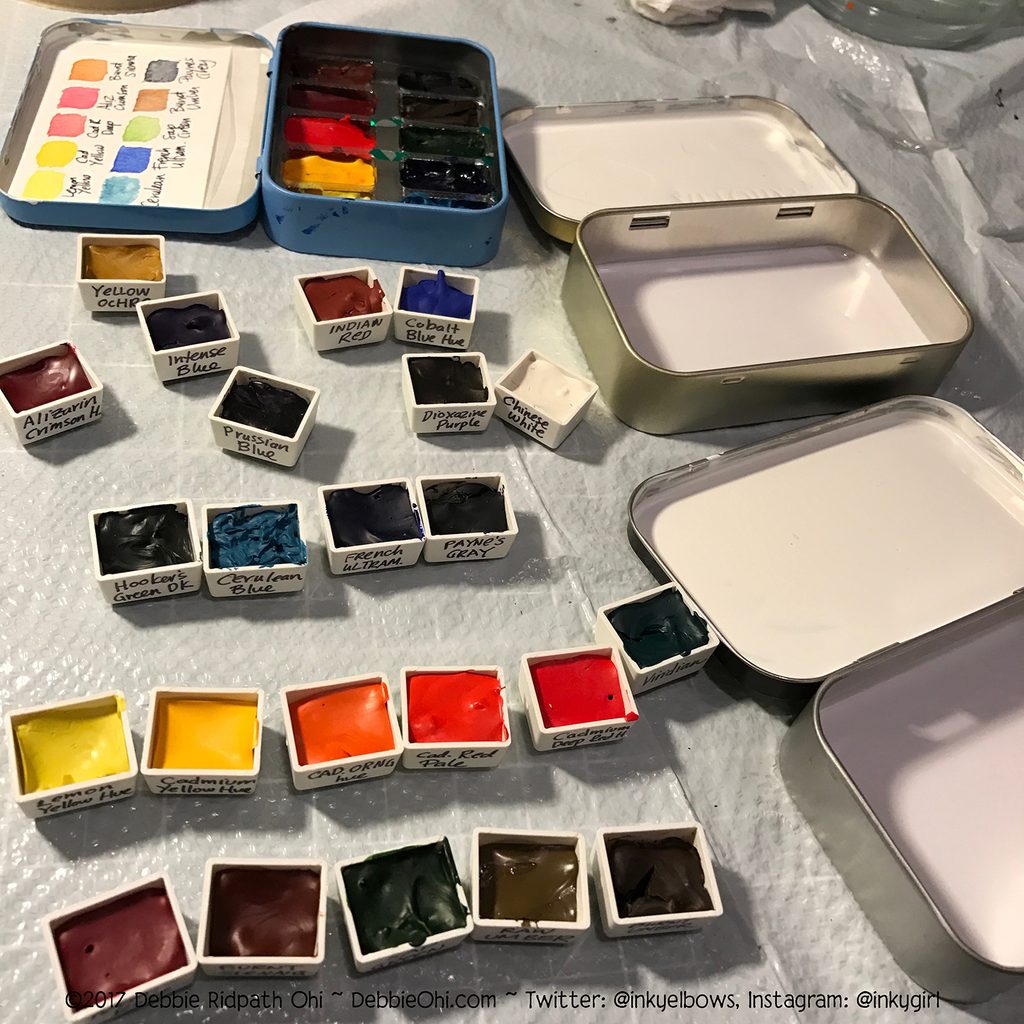 A photo showing two DIY travel watercolor kits in progress, displayed on a work surface next to various paint supplies. One kit features an open metal tin filled with colorful watercolor pans and a handwritten color swatch chart glued inside the lid, while the other shows an open metal tin with its inner lid and base coated in white enamel paint for mixing colors. Copyright 2017 Debbie Ridpath Ohi ~ DebbieOhi.com ~ Twitter: @inkyelbows, Instagram: @inkygirl is visible at the bottom.