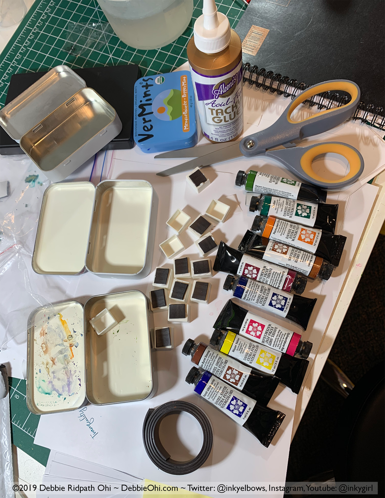 A photo showing supplies for making a DIY travel watercolor kit, including two open metal mint tins, a bottle of glue, a pair of scissors, several tubes of Daniel Smith watercolor paint, a VerMints mint tin, a roll of magnetic tape, and small square magnetic pieces. Copyright 2019 Debbie Ridpath Ohi ~ DebbieOhi.com ~ Twitter: @inkyelbows, Instagram, Youtube: @inkygirl is visible at the bottom.